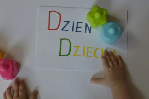 Dziecko pisze "Dzień Dziecka" kolorowymi flamastrami. Obok leżą zabawki.