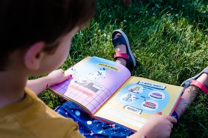 Dziecko na trawie czyta książkę z ilustracjami, ucząc się jak nauczyć dziecko czytać. Na stronach widać pieska i miski z jedzeniem.