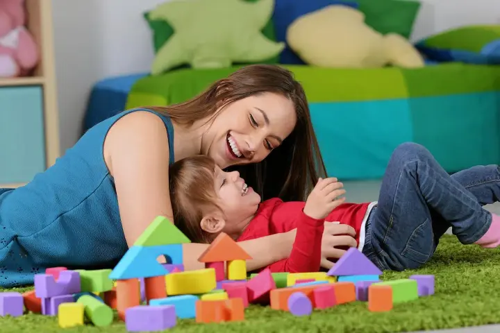 Uśmiechnięta opiekunka bawi się z dzieckiem wśród kolorowych klocków. Zastanawiasz się, ile bierze opiekunka do dziecka za godzinę?